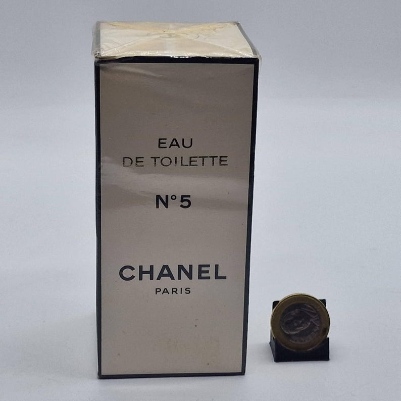 May include: A rectangular perfume box with the text "EAU DE TOILETTE N&deg;5 CHANEL PARIS" in black. The box is beige with a clear plastic wrap. A small coin is next to the box.