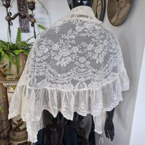 May include: An antique cream lace shawl, featuring detailed floral embroidery and a frilled border. The shawl is displayed on a black mannequin, highlighting the intricate patterns. The openwork design and scalloped edging contribute to its refined aesthetic.
