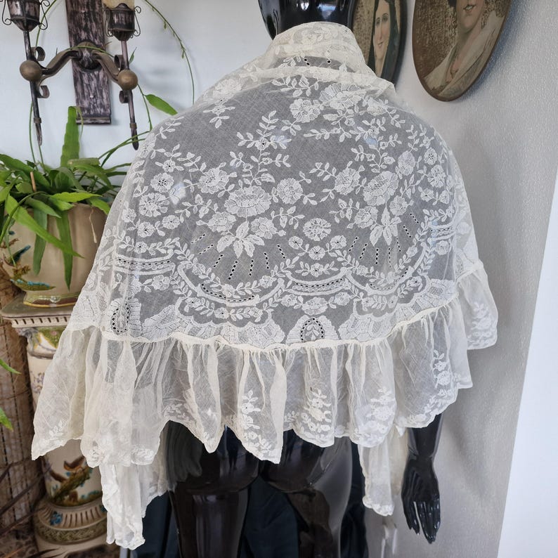 May include: An antique cream lace shawl, featuring detailed floral embroidery and a frilled border. The shawl is displayed on a black mannequin, highlighting the intricate patterns. The openwork design and scalloped edging contribute to its refined aesthetic.