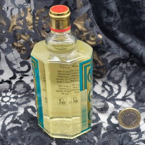 May include: A clear, octagonal perfume bottle with a gold cap and a red top. The bottle has a teal and gold label with text. The liquid inside is yellow. A coin is in the background.
