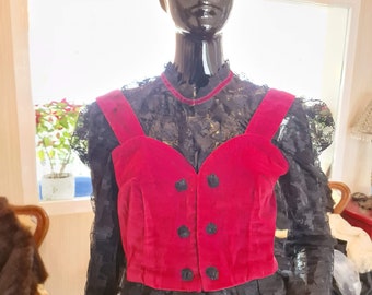 May include: A red velvet cropped vest with black buttons over a black lace top. The vest has a sweetheart neckline and adjustable straps.