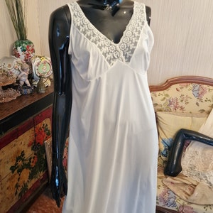 Può includere: Vestito slip bianco con un bordo di pizzo intorno al collo e all'orlo. L'abito è senza maniche e ha una scollatura a V.