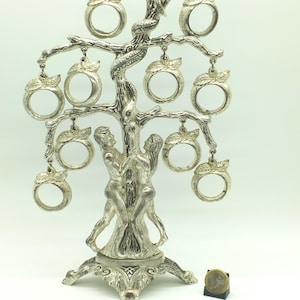Puede incluir: Porta anillos de árbol de metal plateado con una serpiente y dos figuras de pie en el tronco. El árbol tiene nueve anillos colgando de las ramas.