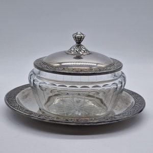 Könnte beinhalten: Eine Butterdose aus Silber und Glas mit einem dekorativen Deckel und einem passenden Teller. Die Dose hat eine klare Glasschale mit einem geschliffenen Glasdesign und einem silbernen Rand. Der Deckel hat einen silbernen Knopf und ein dekoratives silbernes Band um den Rand.