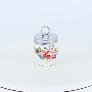 Può includere: Un porta uova in ceramica bianca con un motivo floreale con rose rosa, fiori blu e gialli. Il porta uova ha un coperchio argentato con un manico ad anello.