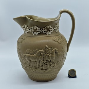Pode incluir: Um jarro de cerâmica castanho com um design em relevo detalhado. O jarro apresenta uma pega e uma borda floral decorativa perto da borda. O relevo retrata figuras a cavalo e num cenário paisagístico. Há uma moeda ao lado do jarro.