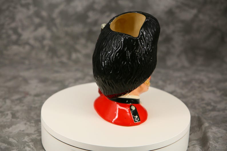 Może przedstawiać: Ceramiczny kubek w kształcie głowy brytyjskiego Gwardzisty Kr&oacute;lewskiego. Kubek ma czarny kapelusz z niedźwiedziej sk&oacute;ry, czerwoną tunikę i szczeg&oacute;łową twarz. Wnętrze kubka jest beżowe. Unikalny przedmiot kolekcjonerski.