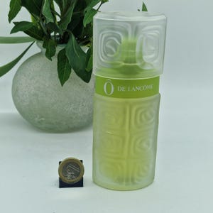Puede incluir: Un frasco de perfume vintage con una tapa transparente de forma cuadrada y un cuerpo verde claro. El frasco tiene un diseño texturizado y el texto "O DE LANCÔME" es visible. Una moneda de un euro está en primer plano.