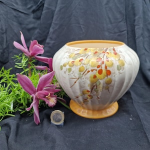 Puede incluir: Un jarrón de cerámica crema, acanalado, con una abertura ancha y una base redonda. El jarrón está decorado con motivos florales amarillos y rojos. Una ramita de flores moradas y follaje verde está junto al jarrón.