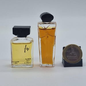 Puede incluir: Dos frascos de perfume vintage con tapones negros y una moneda de un euro. El frasco de la izquierda está etiquetado como "Fiji" de Guy Laroche, París. El otro frasco tiene forma rectangular alta. La moneda está sobre un soporte negro.