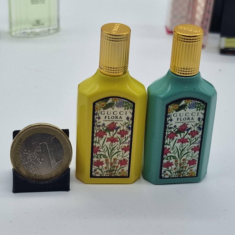 May include: Two small perfume bottles, one yellow and one teal, with gold-colored caps. Each bottle has a label with the words "GUCCI FLORA" and a floral design. A Euro coin is next to the bottles.