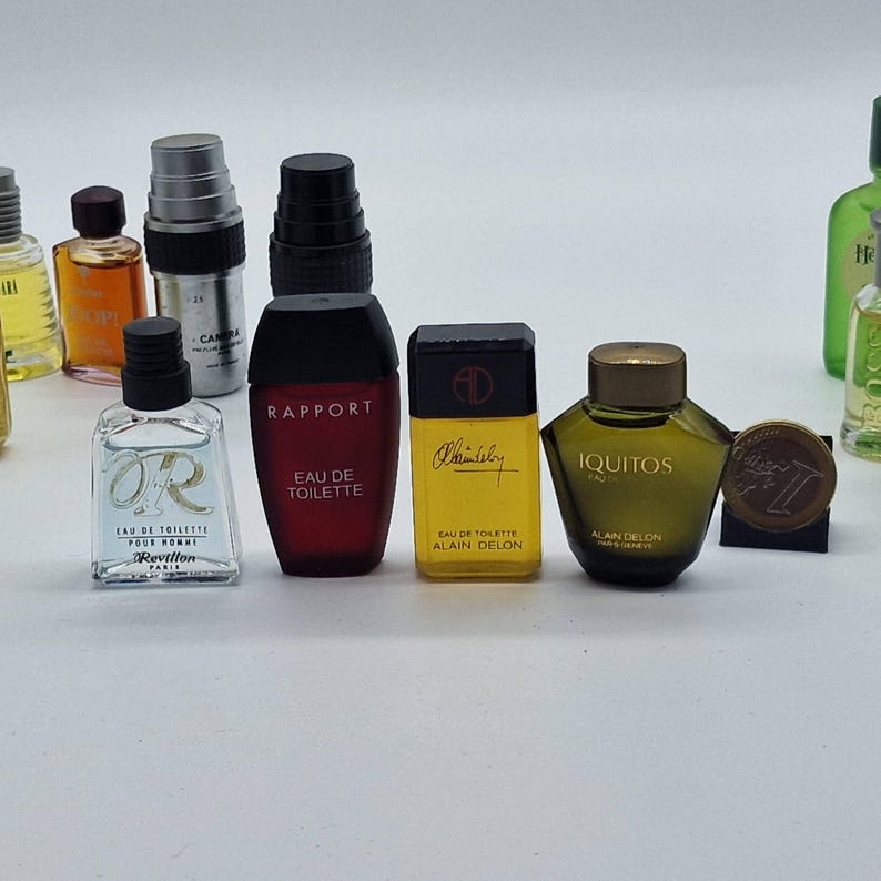 May include: A collection of miniature perfume bottles of various shapes and colors. The bottles include brands like Rapport, Alain Delon, and Revillon. The bottles are arranged on a white surface, showcasing their diverse designs and labels.