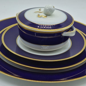 Peut inclure: Un ensemble de trois assiettes en porcelaine bleues à bordure dorée avec un petit bol en porcelaine blanche avec un couvercle et une bordure dorée. Le bol a un bouton blanc sur le couvercle.