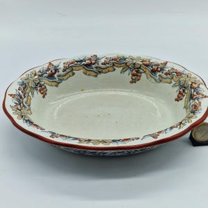 May include: Oval ceramic serving dish with a scalloped rim. The dish features a cream-colored interior and a decorative border with orange berries, yellow ribbons, and blue accents. The rim is a deep red.