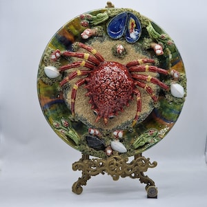 Puede incluir: Un plato decorativo de cerámica con un diseño de cangrejo. El cangrejo es de color rojo oscuro con detalles amarillos, rodeado de arena, conchas y algas marinas. El plato tiene un borde colorido y texturizado y se exhibe en un soporte dorado.
