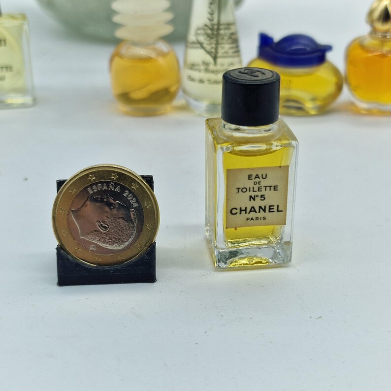 K&ouml;nnte beinhalten: Eine Miniatur-Parf&uuml;mflasche Chanel No. 5 Eau de Toilette mit schwarzem Verschluss und Etikett. Die Flasche ist mit einer gelben Fl&uuml;ssigkeit gef&uuml;llt. Eine Euro-M&uuml;nze befindet sich im Vordergrund, gehalten in einem schwarzen St&auml;nder. Weitere Parf&uuml;mflaschen sind im Hintergrund.