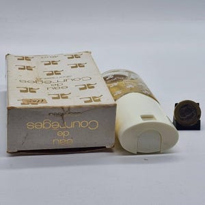 May include: A vintage Courr&egrave;ges Eau de Toilette atomizer and box. The box is beige with gold lettering and a repeating logo pattern. The atomizer is white and clear, with a cylindrical shape. A coin is also present.