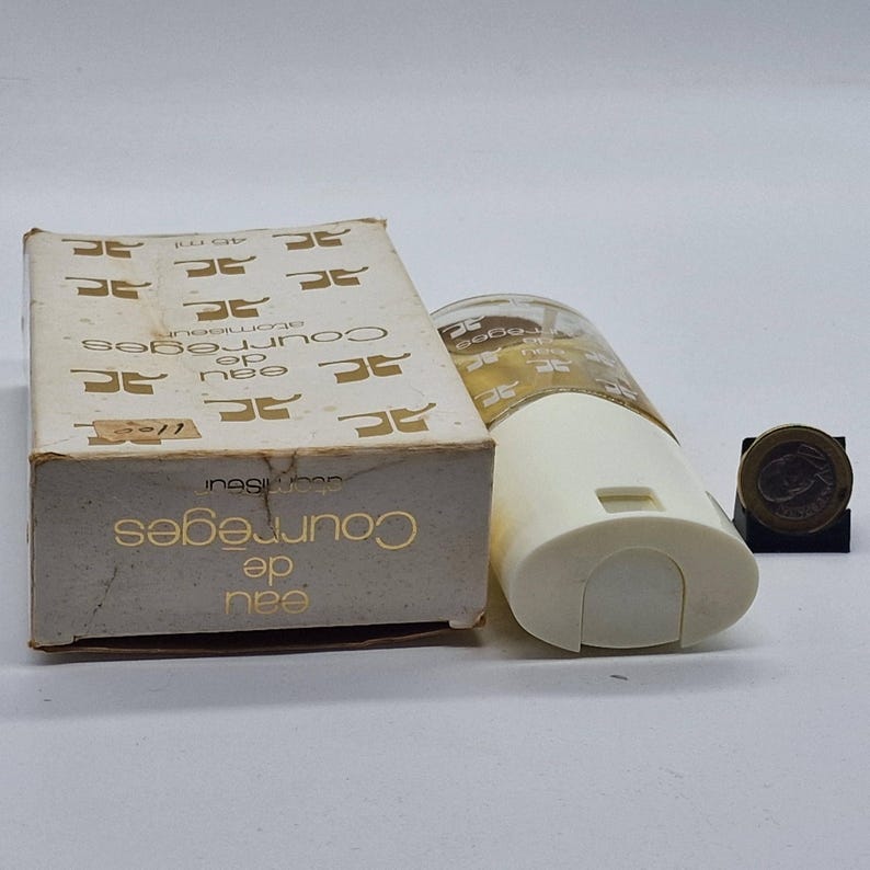 May include: A vintage Courr&egrave;ges Eau de Toilette atomizer and box. The box is beige with gold lettering and a repeating logo pattern. The atomizer is white and clear, with a cylindrical shape. A coin is also present.