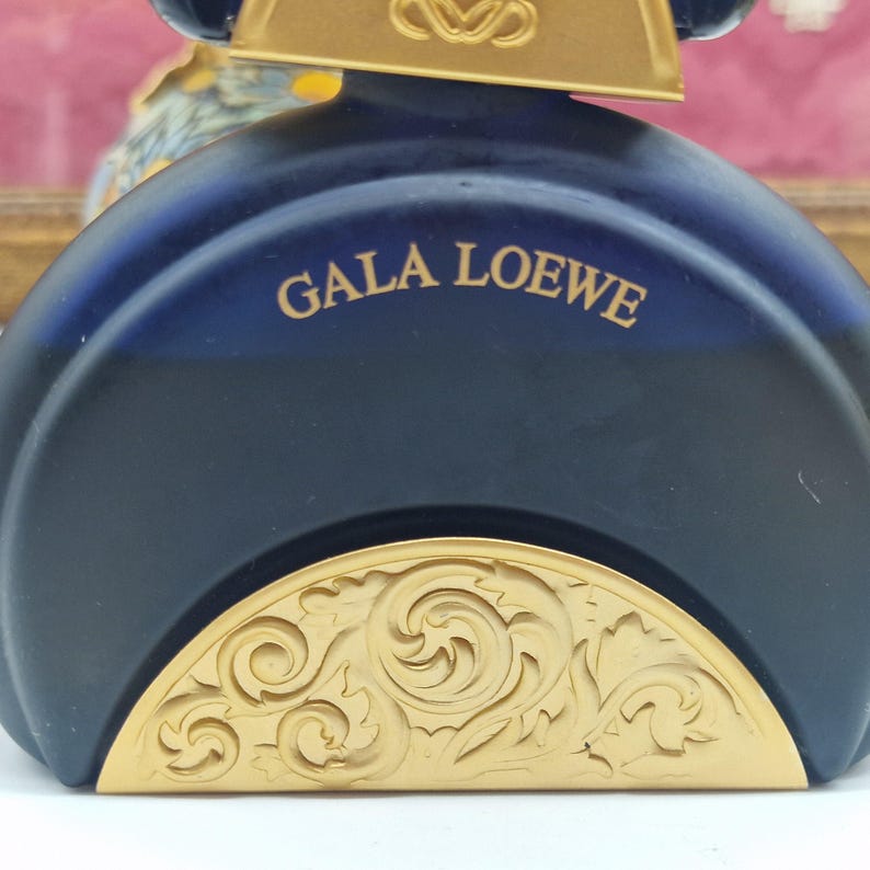 May include: A dark blue perfume bottle with the words "GALA LOEWE" in gold lettering. The bottom of the bottle features a gold, ornate, floral design. The top of the bottle is gold with a decorative design.