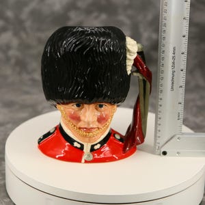 Może przedstawiać: Ceramiczny kubek w kształcie brytyjskiego Gwardzisty Kr&oacute;lewskiego. Kubek ma czarny kapelusz z niedźwiedziej sk&oacute;ry, czerwoną kurtkę i szczeg&oacute;łową twarz. Uchwyt jest czerwony i zielony. Linijka pokazuje wysokość kubka w cm.