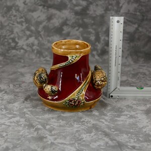 Peut inclure: Un vase en céramique rouge foncé avec une finition brillante, présentant des accents décoratifs en relief de couleur beige, incluant des formes sculptées rappelant des coquillages et des détails floraux. Une règle en métal en L indique 10 cm.