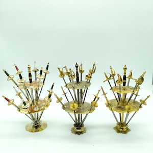 Peut inclure: Trois supports en métal doré avec une base circulaire et un dessus décoratif. Chaque support contient une collection d'épées miniatures avec des poignées dorées et noires. Les épées sont disposées en cercle sur le support.