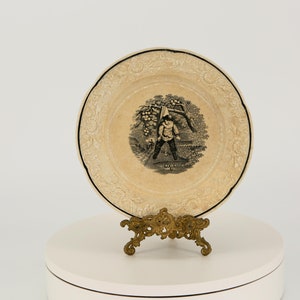 May include: A cream-coloured decorative plate with a black rim and a detailed floral border. The centre features a black and white illustration of a person swinging a golf club. The plate is displayed on a decorative gold-coloured stand.