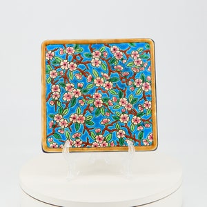Puede incluir: Un azulejo de cerámica cuadrado con fondo azul y un patrón floral de flores rosas y hojas verdes. El azulejo tiene un borde dorado.