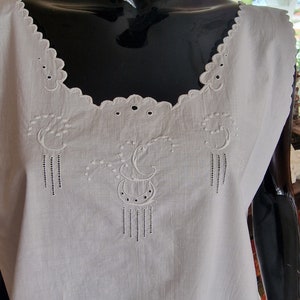May include: White sleeveless cotton top with delicate embroidered details around the neckline and a floral design in the center.