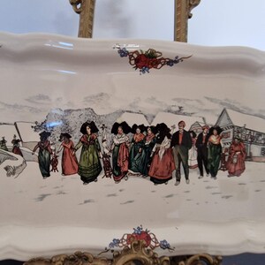 Può includere: Un vassoio da portata in ceramica bianca con una scena invernale che presenta un gruppo di persone in abiti tradizionali che camminano attraverso un villaggio innevato. Il vassoio ha un bordo decorativo con rose rosse e fiori blu.