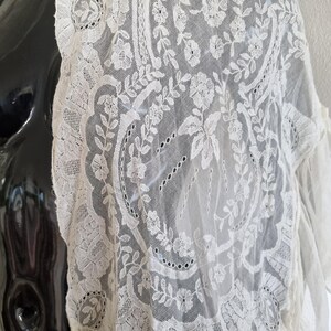 May include: Cream-coloured lace fabric with floral and leaf patterns. The delicate lace is draped over a black mannequin, showcasing the intricate details of the design. The lace features a scalloped edge and a variety of floral motifs.