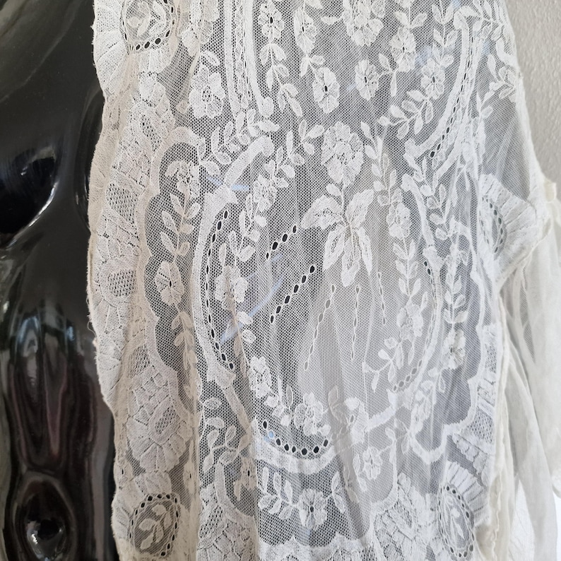 May include: Cream-coloured lace fabric with floral and leaf patterns. The delicate lace is draped over a black mannequin, showcasing the intricate details of the design. The lace features a scalloped edge and a variety of floral motifs.