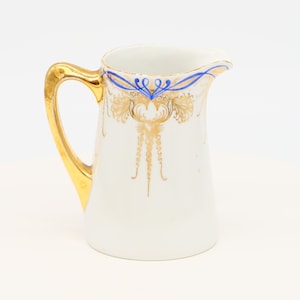 Puede incluir: Jarra de cerámica blanca con asa dorada y detalles florales dorados y azules. La jarra tiene un pico curvo y un asa ancha de color dorado. El diseño incluye patrones florales dorados y detalles azules cerca del borde.