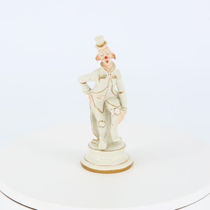 Könnte beinhalten: Eine weiße Porzellanfigur eines Clowns mit einem Zylinder, einer roten Nase und goldenen Akzenten. Der Clown steht mit einem gebeugten Bein und hält eine Blume in der Hand.