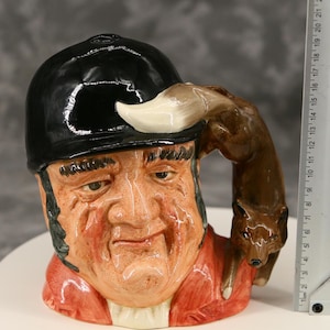 Peut inclure: Mug en céramique représentant une tête d'homme avec un casque d'équitation noir et un manteau rouge. La anse est formée par un renard brun. Le mug mesure environ 15 cm de hauteur et présente des détails peints.