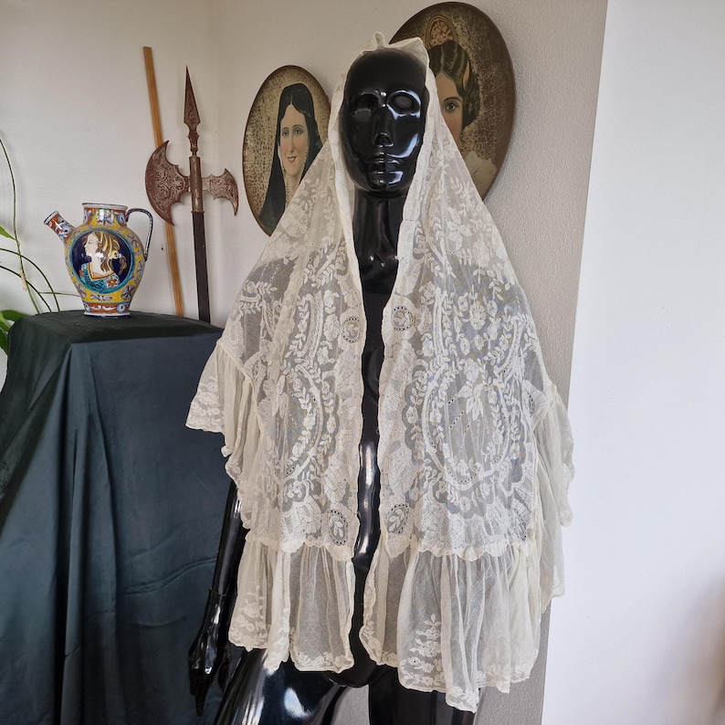 May include: Cream-coloured lace shawl with intricate floral and scroll patterns. The shawl has a ruffled trim and is displayed on a black mannequin. The background includes antique portraits and decorative items, suggesting a vintage style.