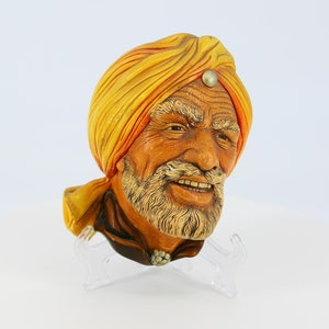 Peut inclure: Une plaque murale en céramique représentant la tête d'un homme portant un turban jaune avec une bordure orange. L'homme a une barbe et une moustache blanches et sourit.