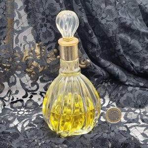 May include: A vintage perfume bottle with a clear, ribbed glass body and a decorative, clear stopper. The bottle contains a yellow liquid and has a gold-colored metal neck. The background is a black lace fabric.