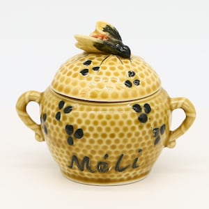 May include: A ceramic honey pot with a bee on the lid. The pot is decorated with a honeycomb pattern and the word "Méli" is written on the front.