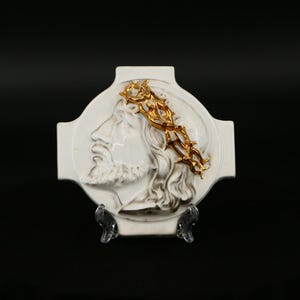 May include: White ceramic wall plaque featuring a raised relief of Jesus's face with a gold crown of thorns. The plaque is shaped like a cross and has a stand.