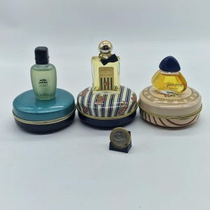 Puede incluir: Tres botellas de perfume en miniatura sobre latas decorativas. Las botellas tienen diferentes formas y colores, con etiquetas. Las latas son redondas y presentan varios diseños, incluyendo rayas y motivos florales. Una moneda está en primer plano.
