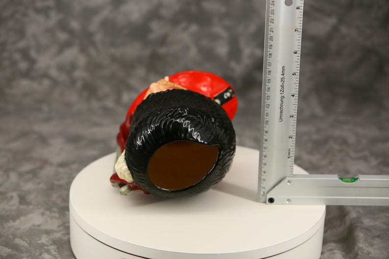 Może przedstawiać: Ceramiczna figurka gnoma z czarną brodą, czerwonym kapeluszem i czerwoną kurtką. Gnom leży na brzuchu. Obok gnoma znajduje się linijka, pokazująca wymiary w centymetrach.