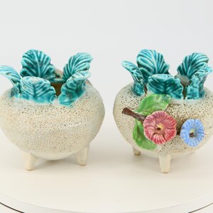 Puede incluir: Dos pequeños maceteros de cerámica blanca con tres patas cada uno. Los maceteros están decorados con hojas verdes y flores de colores.