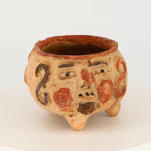 Op de afbeelding: Een kleine terracotta pot met een gezicht erop geschilderd. Het gezicht heeft rode en zwarte details en de pot heeft drie poten.