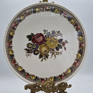 May include: A white ceramic plate with a gold trim and a floral design. The plate features a bouquet of colourful flowers with green leaves and a brown background.