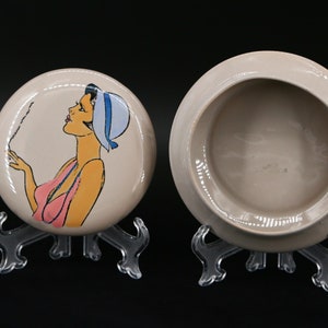 May include: A vintage ceramic trinket box with a lid. The lid features a hand-painted illustration of a woman in a pink swimsuit and a blue hat. The woman is holding a cigarette and blowing smoke.