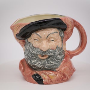 Op de afbeelding: Een keramische kan in de vorm van een mannenhoofd met een rood-zwarte hoed, een grijze baard en een rood-gele robe.