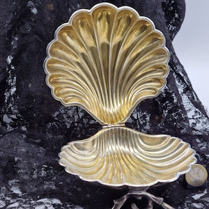 Pode incluir: Uma caixa de joias de prata em forma de concha, com uma tampa articulada. A caixa está aberta, revelando o interior dourado. A caixa está sentada em um tecido de renda preto.