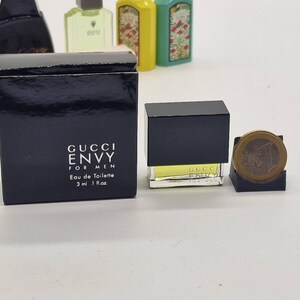May include: A miniature Gucci Envy for Men Eau de Toilette bottle and box. The black box has white text, and the bottle is a small, rectangular shape with a black cap. The bottle contains a yellow liquid. A coin is next to the bottle.