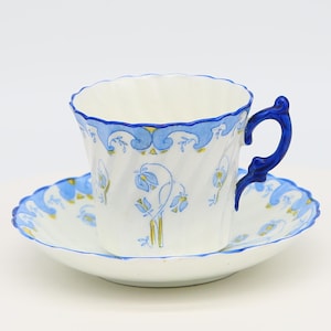 Könnte beinhalten: Eine weiße Teetasse und Untertasse mit einem blauen und goldenen Blumendesign. Die Tasse hat einen blauen Henkel und die Untertasse einen gewellten Rand.